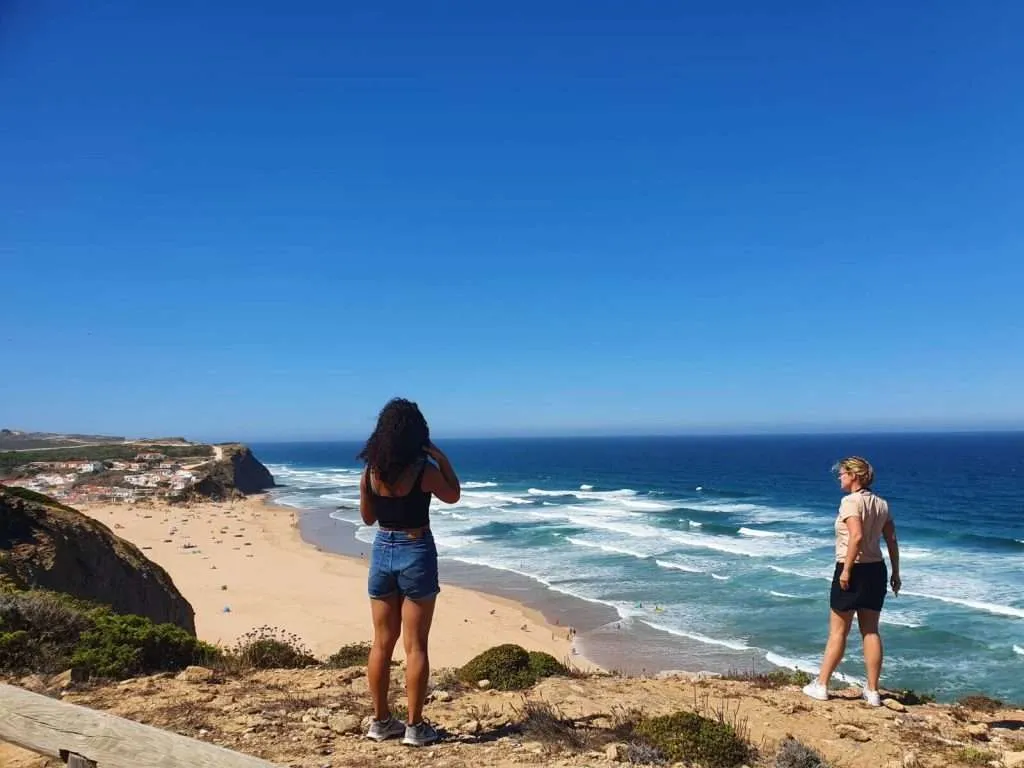 Two women stand on a cliffside path overlooking the vast Atlantic and sandy beach far below, with dramatic waves and a coastal town visible in the distance – one of my favourite things to do in sagres portugal