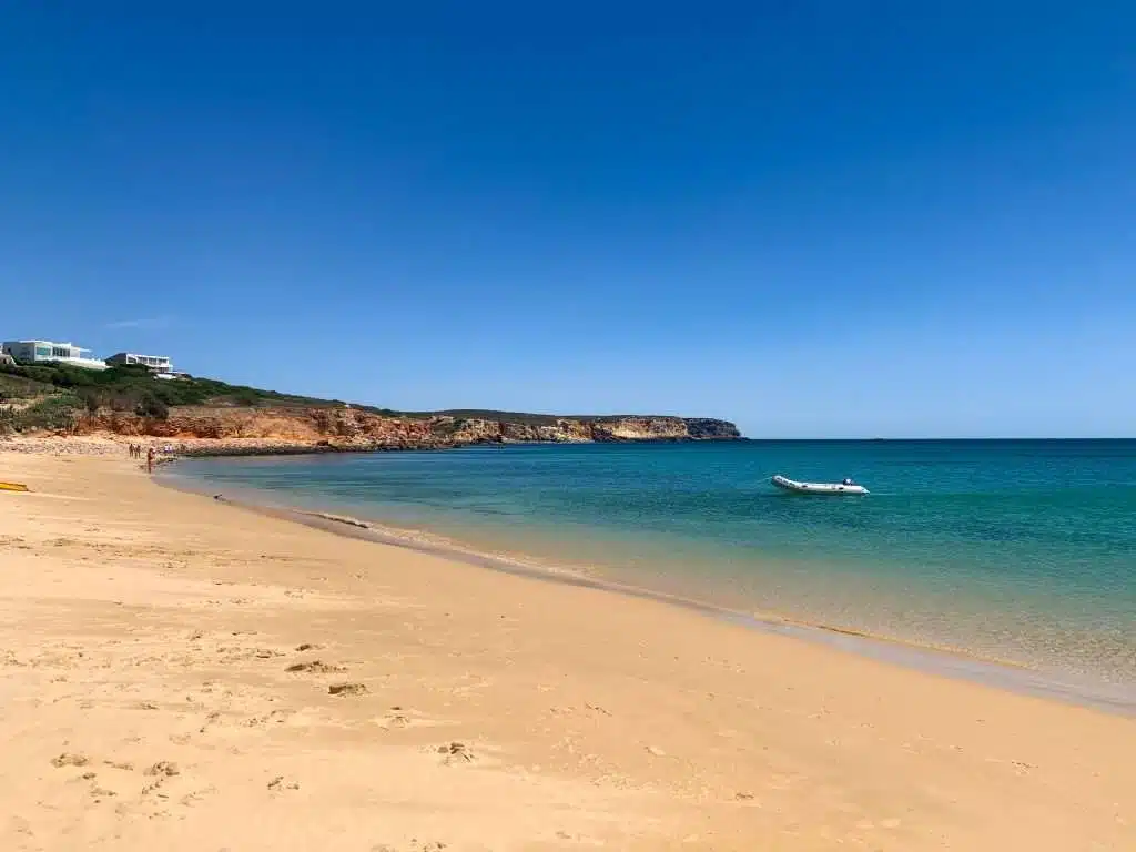 The image shows Praia Do Martinhal with golden sand, gentle turquoise waves, and a small inflatable boat floating offshore; a few beachgoers walk in the distance beneath a clear blue sky. Praia Do Martinhal is one of the best things to do in Sagres, Portugal.