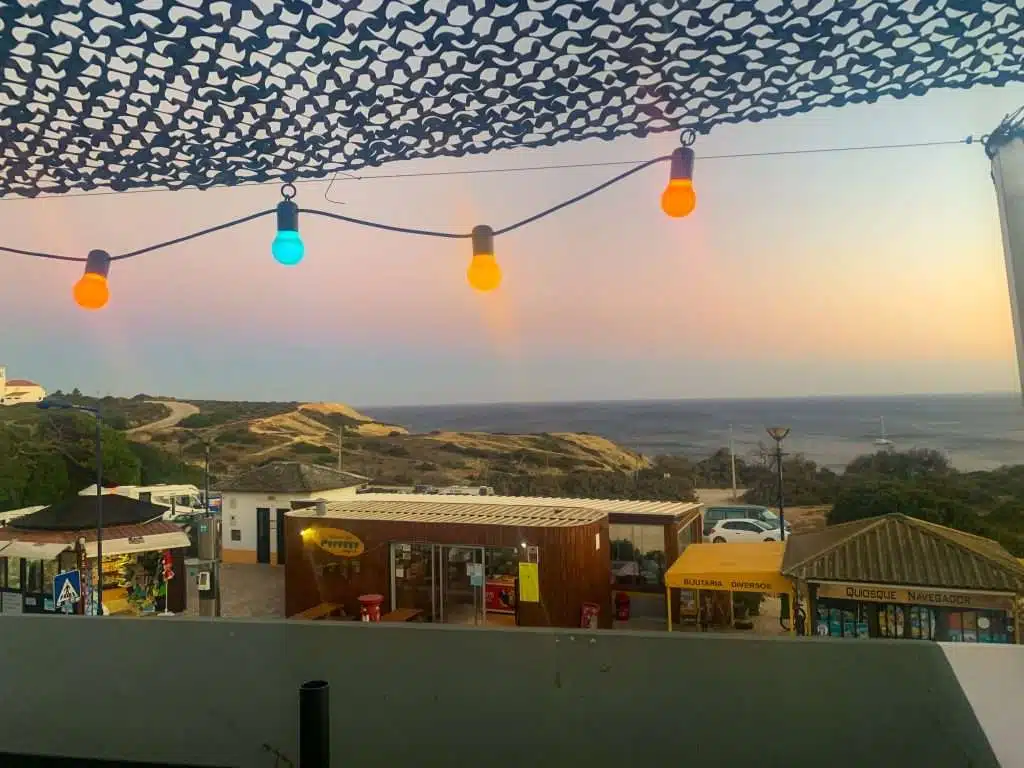 Colorful string lights hang under a netted canopy as the sun sets over a scenic coastal village, with small food kiosks and the ocean beyond rolling hills in the background.