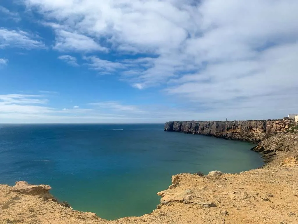 Panoramic view of the coastline featuring sheer, rugged cliffs that plunge into calm turquoise waters under a partly cloudy sky.