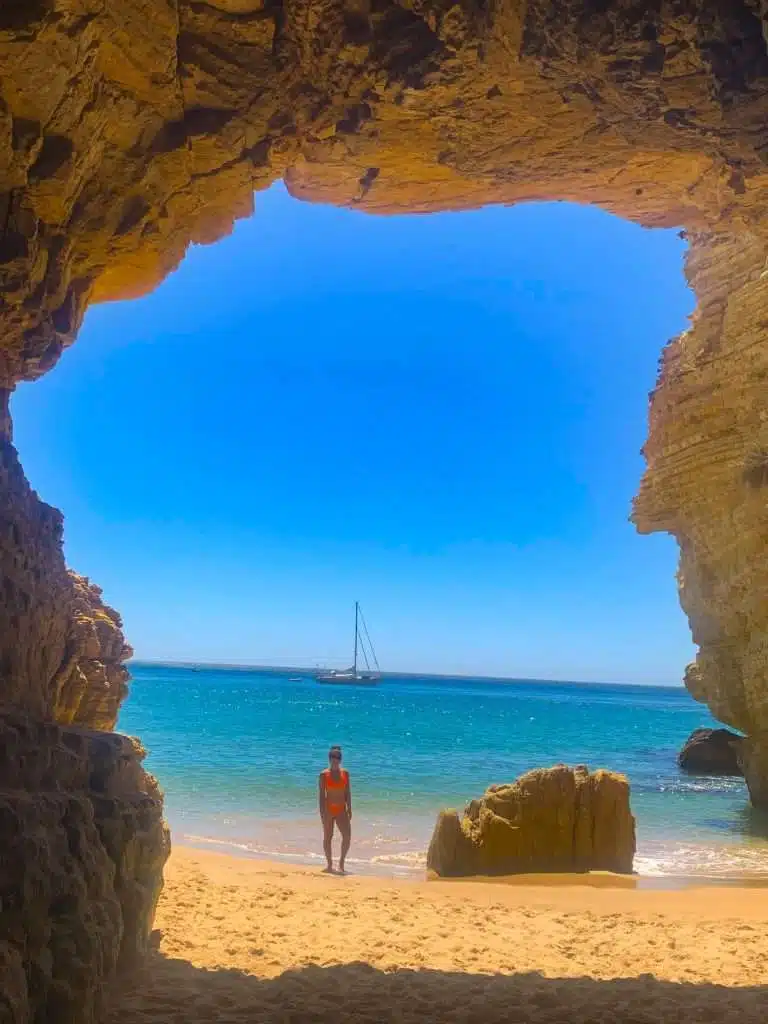 A person in a red swimsuit stands on a sandy beach, framed by the opening of a rocky cave—a perfect scene among the many things to do in Sagres—with turquoise water and a distant sailboat under a bright blue sky.