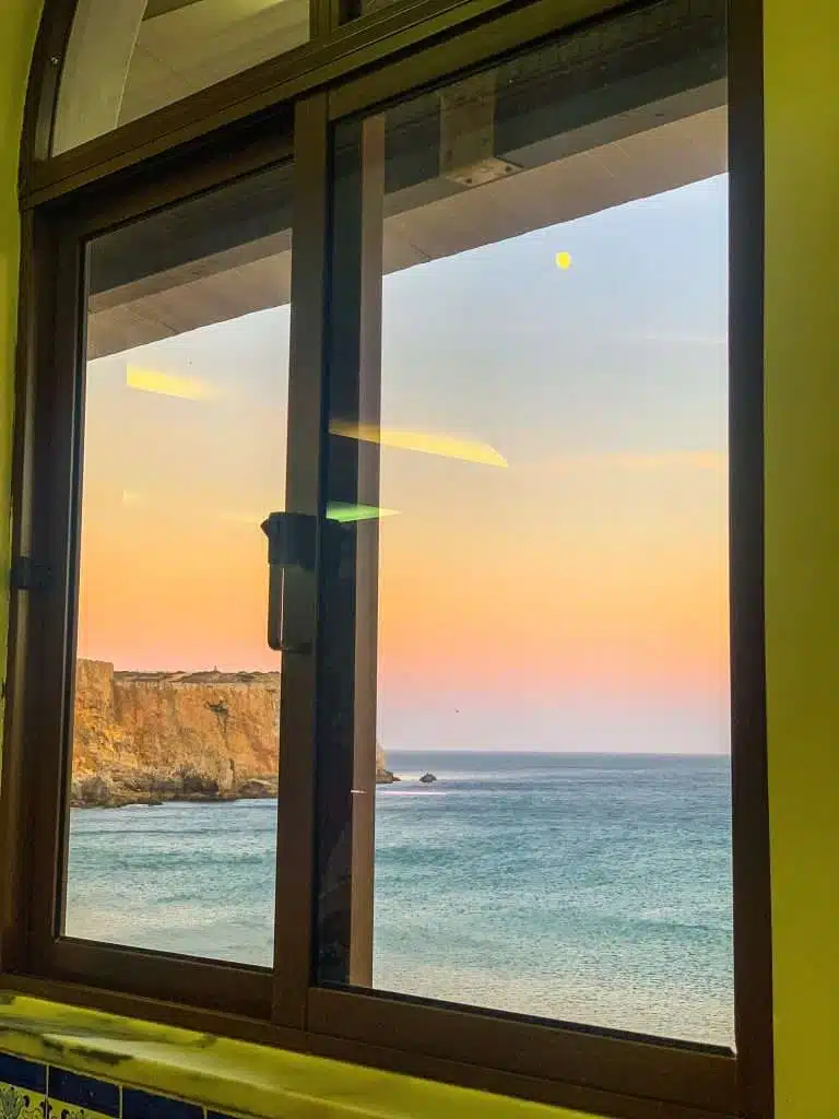 Framed view through a window looking out at the ocean and cliffs of sagres portugal during sunset, with pastel hues in the sky and the moon faintly visible above.