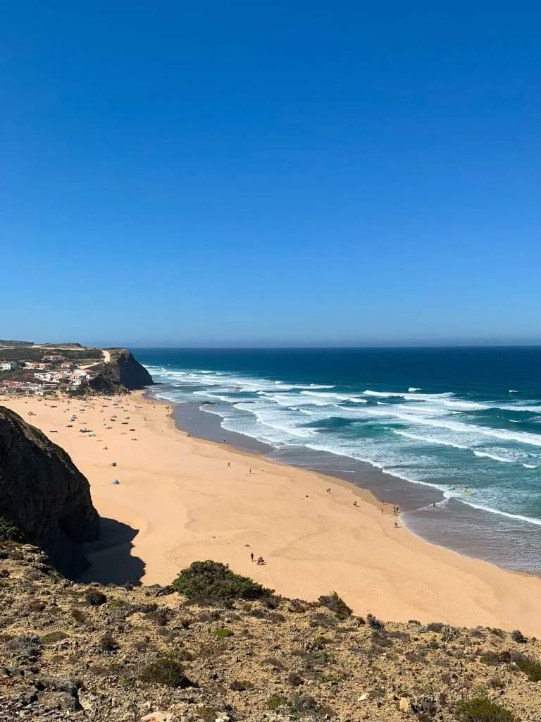 Expansive coastline view from a cliff overlooking a sandy beach with rolling surf and scattered beach tents, with a whitewashed village perched at the base of rugged cliffs.