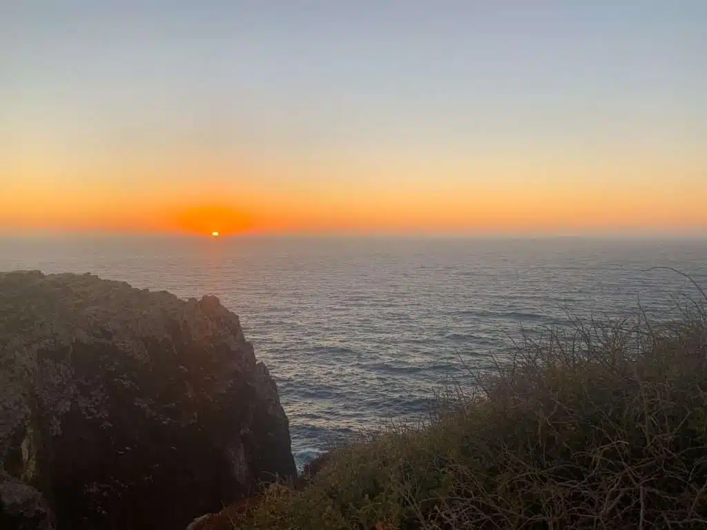 The sun sets over a calm ocean in Sagres, casting an orange glow across the sky. Rocky cliffs and some dry brush are visible in the foreground—just one of many breathtaking things to do in Sagres as gentle waves stretch into the horizon.