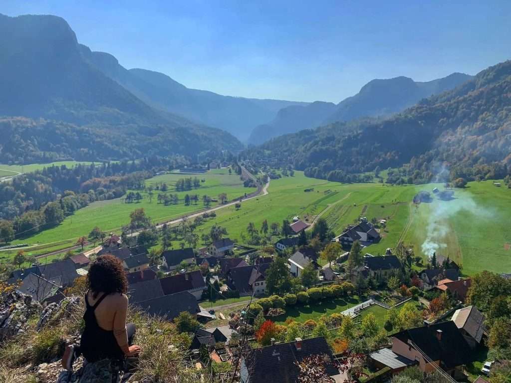 A person with curly hair sits on a rocky ledge overlooking a picturesque valley. Below are a cluster of houses, green fields, and trails of smoke. Mountains rise in the background under a clear blue sky.