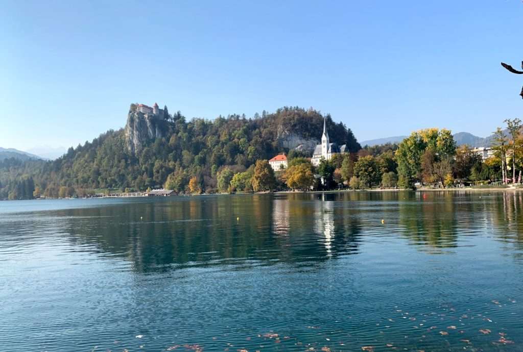 A serene lake with clear, calm water reflecting trees and buildings. A small island with a white church is in the center, surrounded by lush greenery. In the background, a castle sits atop a rocky hill under a clear blue sky.