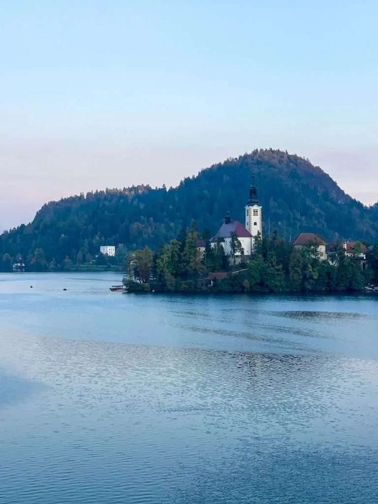 A serene lakeside scene with a small island featuring a church and tower, surrounded by trees. The backdrop includes a hill covered in greenery, under a soft blue sky, reflecting on the calm water.