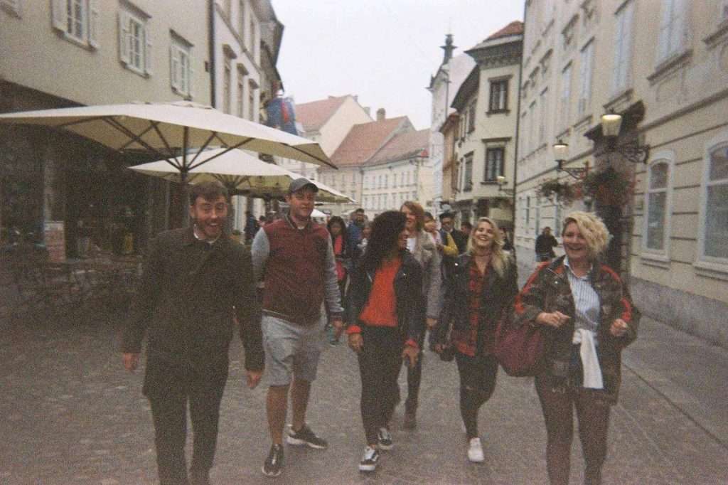 A group of people walking down a narrow, cobblestone street in a historic area with old-style buildings. Some are smiling or talking, and there are large outdoor umbrellas on the sidewalk. The scene has a casual, candid atmosphere.