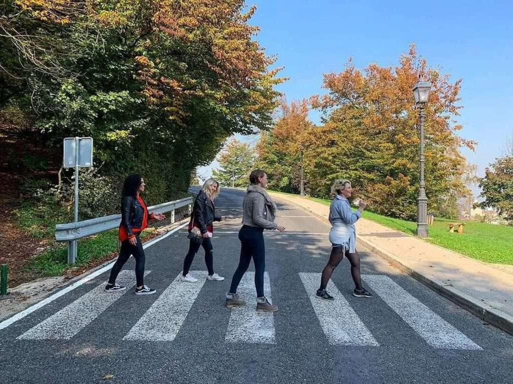 Four women walk across a zebra crossing resembling the famous Abbey Road photo. The street is lined with trees displaying autumn colors. The sky is clear and blue, and a streetlamp is visible on the right.