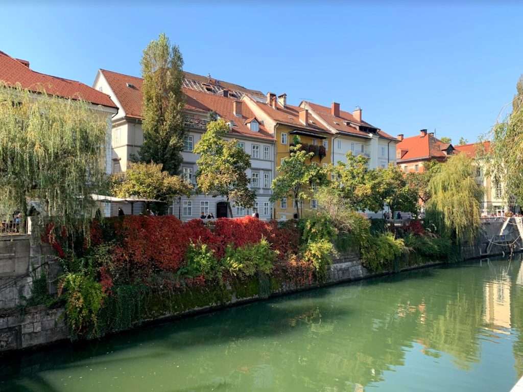 A serene river view with green water lined by trees and plants draped in red and green foliage. Behind, charming houses with red-tiled roofs and white facades sit under a clear blue sky, creating a picturesque scene.