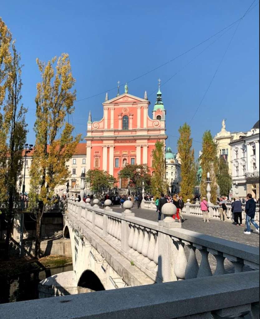 A sunny day view of the Triple Bridge in Ljubljana, Slovenia, featuring people walking across. In the background is the pink facade of the Franciscan Church of the Annunciation surrounded by trees with autumn foliage.