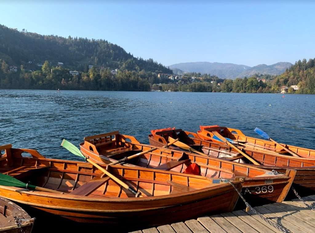 Wooden rowboats are moored to a dock, with calm blue water and forested hills in the background under a clear sky.