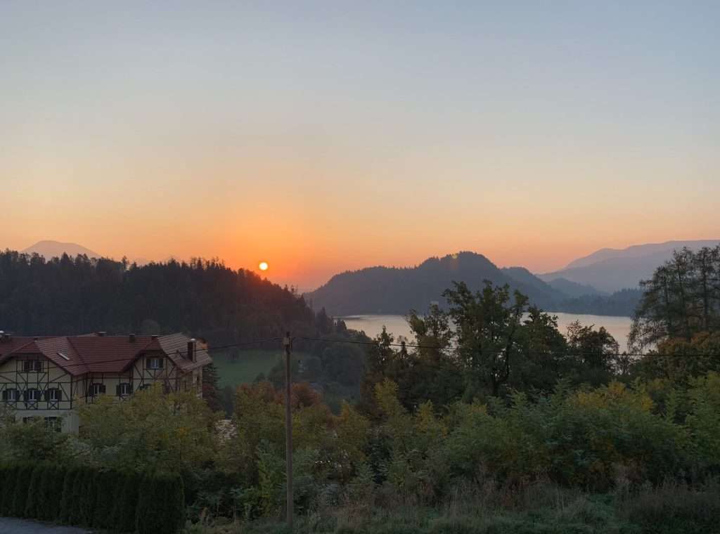 A picturesque sunrise over a serene landscape with misty hills, a valley filled with trees, and a house with a red roof in the foreground. The sky transitions from orange to blue as the sun rises.