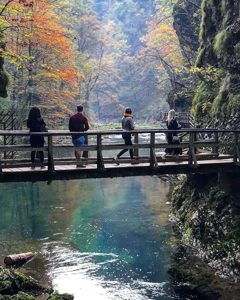 Four people walk across a wooden bridge over a clear, reflective river. Surrounding them are rocky cliffs and vibrant autumn foliage, with a mix of green, orange, and yellow leaves. The scene is serene and picturesque.