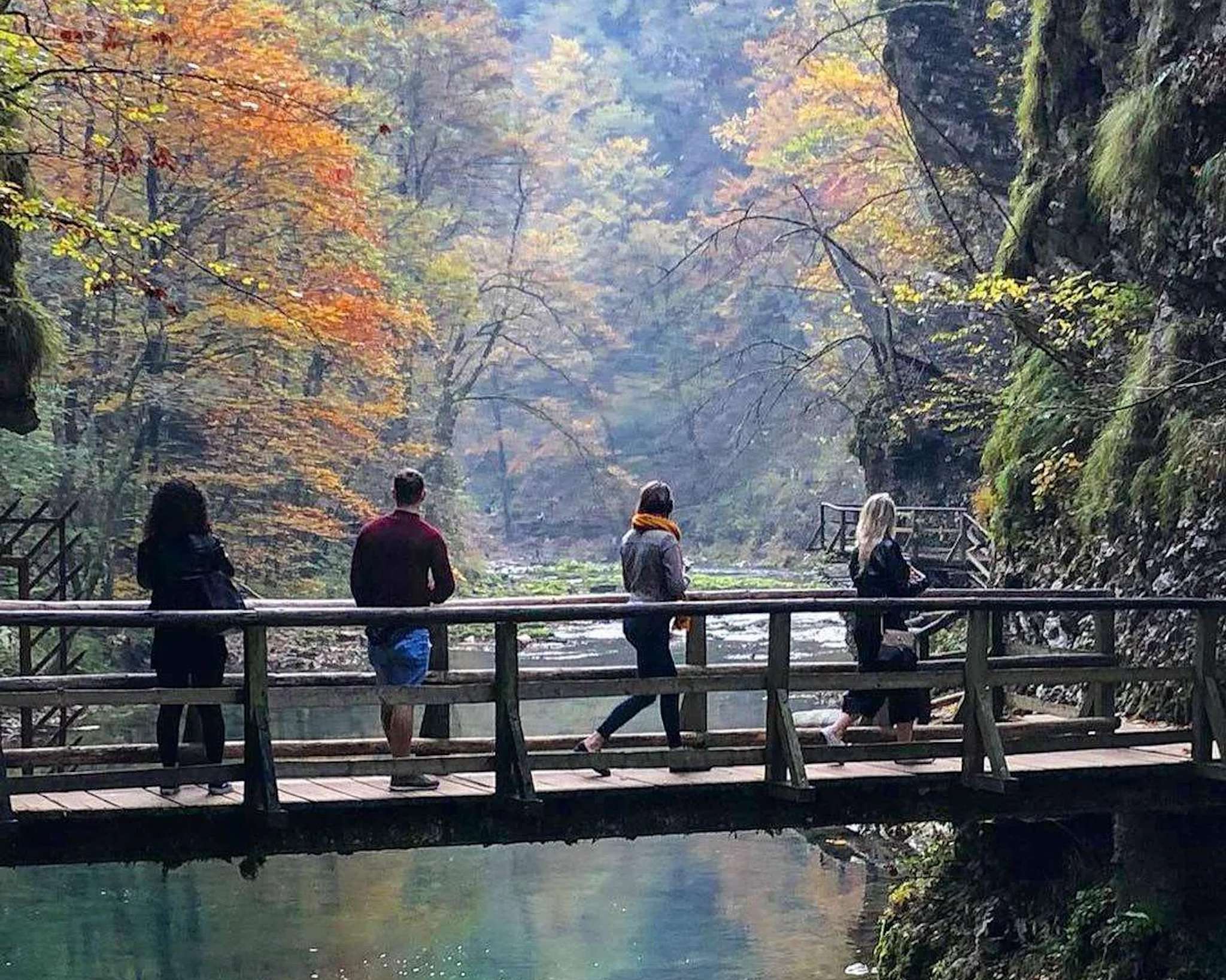 Four people stroll across a wooden bridge spanning a peaceful river in a forested area during autumn, reminiscent of the serene journey from Ljubljana to Lake Bled. The surrounding trees display vibrant fall colors, with shades of orange, yellow, and green. The scene is calm and picturesque.