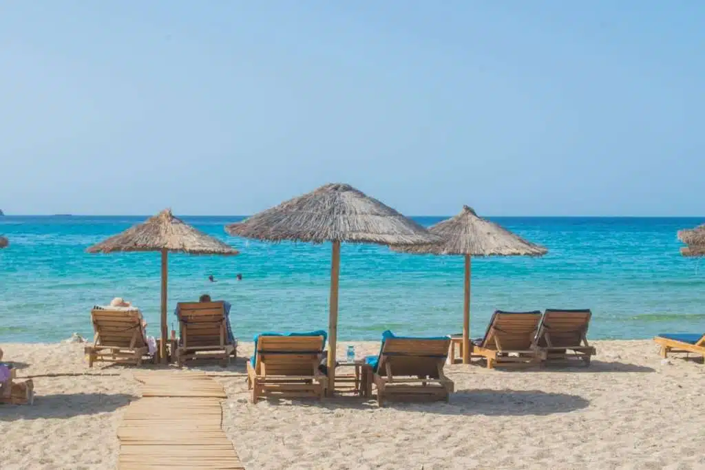 Beach scene with straw umbrellas and lounge chairs on the sand. A wooden walkway leads to the water's edge. Two people are swimming in the clear blue sea under a clear sky, perfectly capturing one of the serene things to do in Paros.