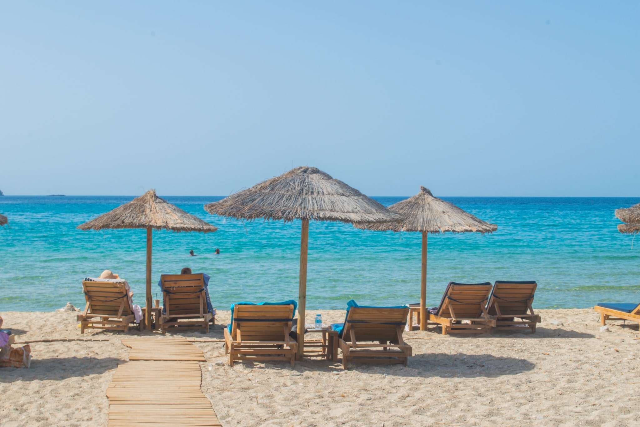 Beach scene with straw umbrellas and lounge chairs on the sand. A wooden walkway leads to the water's edge. Two people are swimming in the clear blue sea under a clear sky, perfectly capturing one of the serene things to do in Paros.