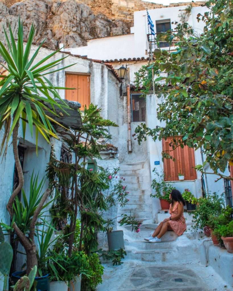 A woman in a coral dress sits on steps in a charming, narrow alleyway in the village of Anafiótika surrounded by lush greenery, colorful potted plants, and white-washed buildings—a perfect glimpse into one of the many things to do in Athens. A rocky hill rises in the background.