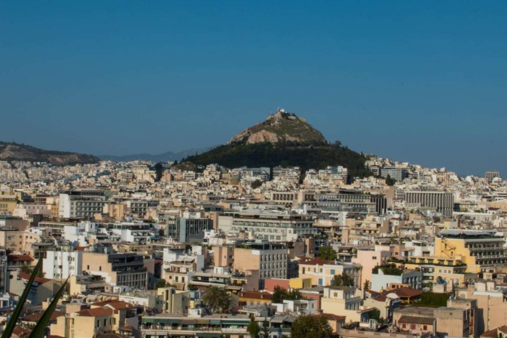 A scenic view of a dense Athens with numerous buildings under a clear blue sky. In the background, a hill covered in green trees rises prominently, with a structure visible at its peak. This panorama could be part of your 5-Day Greece Itinerary, where urban charm and natural beauty coexist harmoniously.