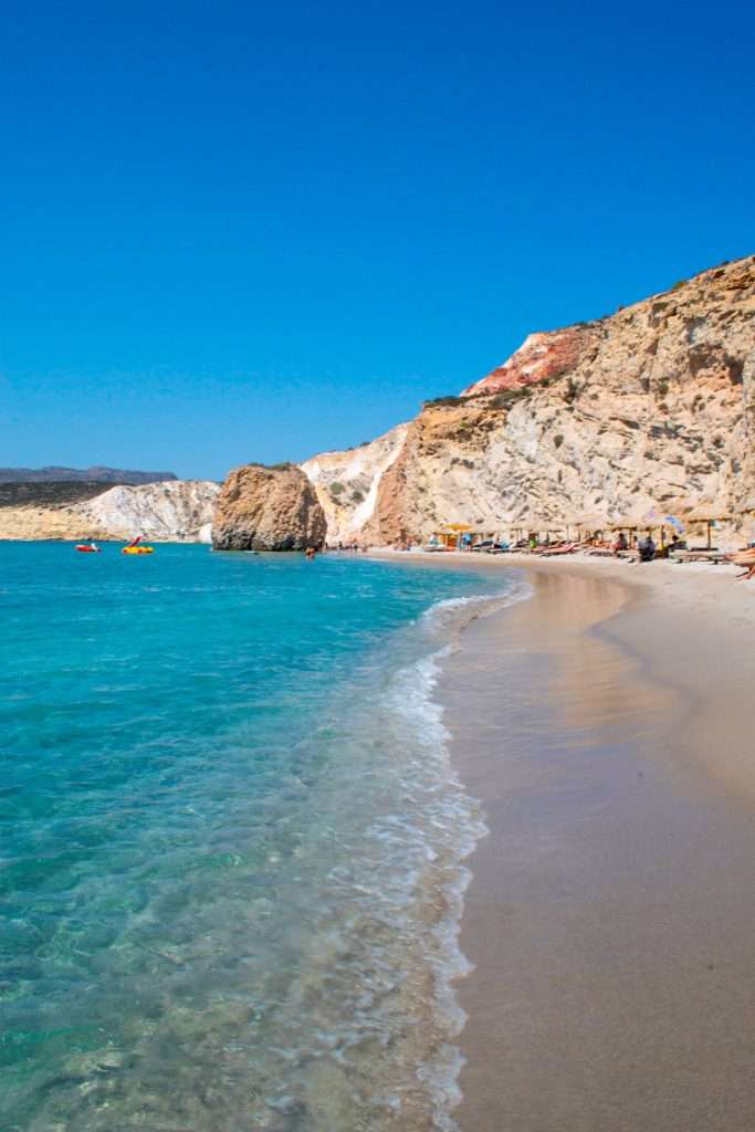 A picturesque image of Fyriplaka Beach. It has clear turquoise water gently lapping onto a sandy shore. The coastline is lined with rugged, sunlit cliffs, and people are visible enjoying the beach and ocean activities—a perfect glimpse of things to do in Milos. The sky is a clear, vibrant blue.