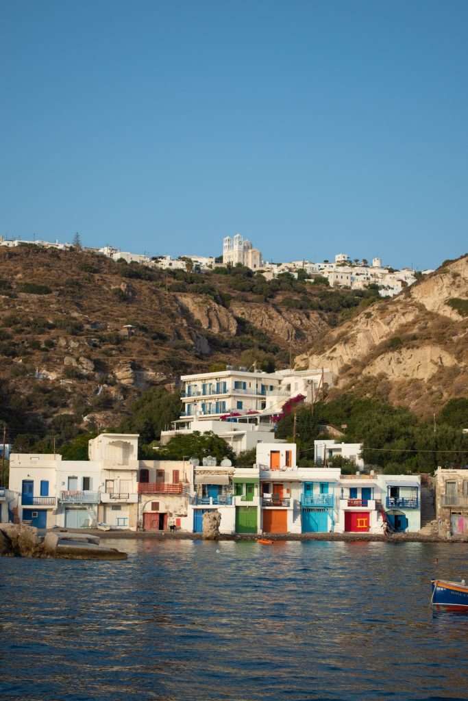 A coastal scene featuring a row of colorful buildings at the water's edge, with blue, green, red, and orange doors. Behind them is a hill with additional white buildings, topped by a church with twin towers. This picturesque view is one of the many things to do in Milos under a clear blue sky.