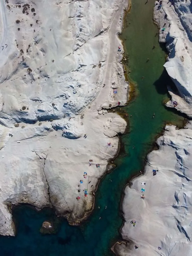 Aerial view of white rock formations and turquoise water at Sarakiniko Beach, Milos, Greece. People are sunbathing and swimming, scattered across the rocky landscape. The contrast between the white rocks and vibrant water creates a striking visual scene, making it a must on the list of things to do in Milos.