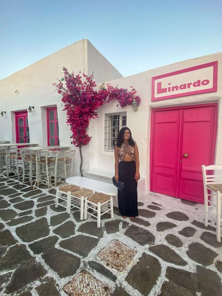 A woman stands smiling in front of a white building with bright pink doors and windows. The building features a sign that reads "Linardo" in pink letters. There's a tall pink-flowering tree beside her, and the ground is paved with stone tiles in white mortar—one of many things to do in Paros.