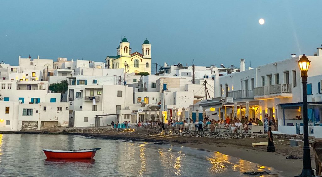 A serene coastal view at dusk shows white buildings with blue accents, a church with two towers lit up in the background, and a full moon in the sky. People are dining at waterfront restaurants while a red boat floats on the calm water in the foreground—a perfect snapshot of things to do in Paros.