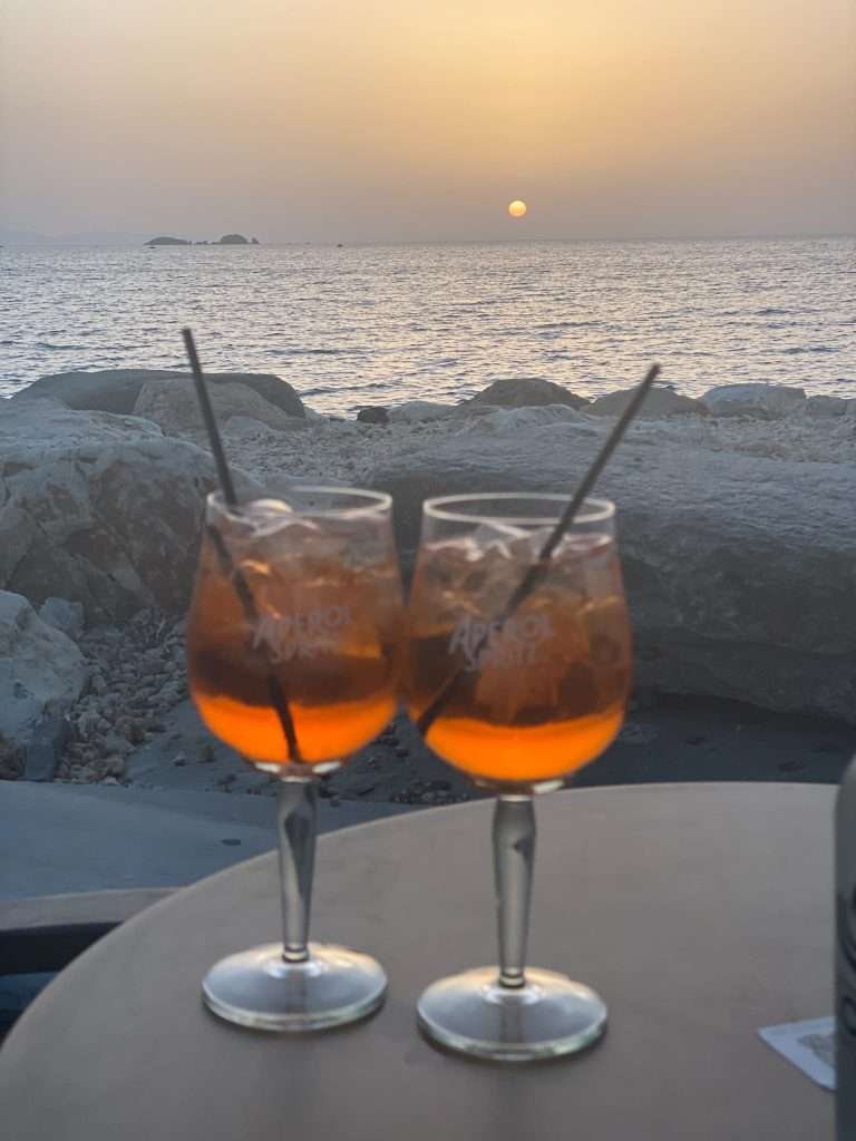 Two glasses of Aperol Spritz with straws are placed on a table overlooking a serene seascape in Paros. As the sun sets on the horizon, casting a warm, golden hue across the sky and water, consider adding this picturesque moment to your list of things to do in Paros. Large rocks form a barrier between the table and the sea.