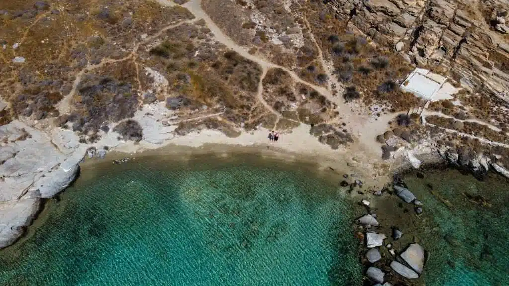 Aerial view of a rocky coastline with clear turquoise waters gently meeting a narrow sandy beach. Small paths traverse the rocky terrain, leading to a white building near the right edge of the image. Among the things to do in Paros, exploring this serene landscape is a must. Sparse vegetation dots the area.