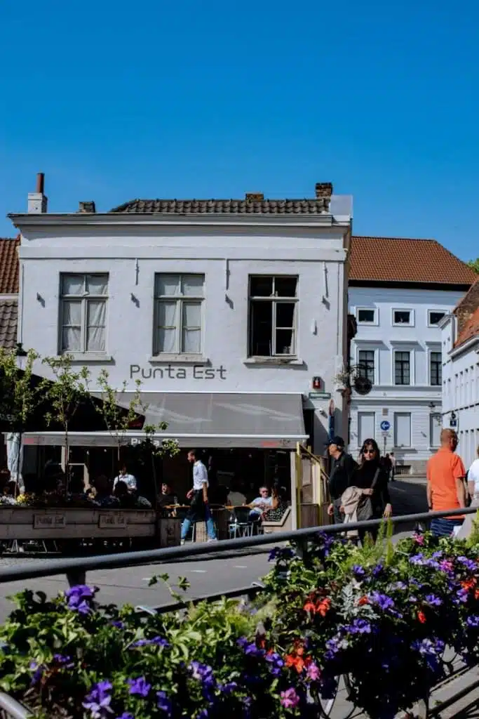 A bustling European street scene in Bruges with people walking past a two-story white building labeled "Punta Est." The foreground features vibrant purple and pink flowers. The sky is clear and blue.