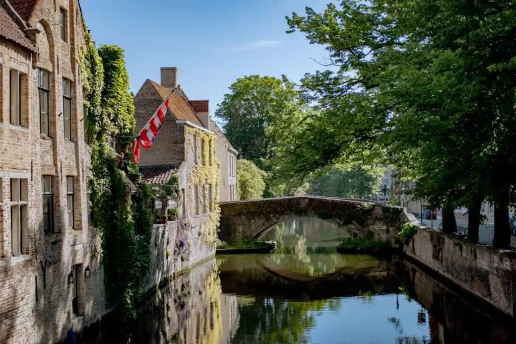 A scenic canal in Bruges, Belgium, flanked by brick buildings covered with ivy. A small stone bridge arches over the tranquil water, which reflects the lush greenery and architecture. A red and white flag flies from one building.