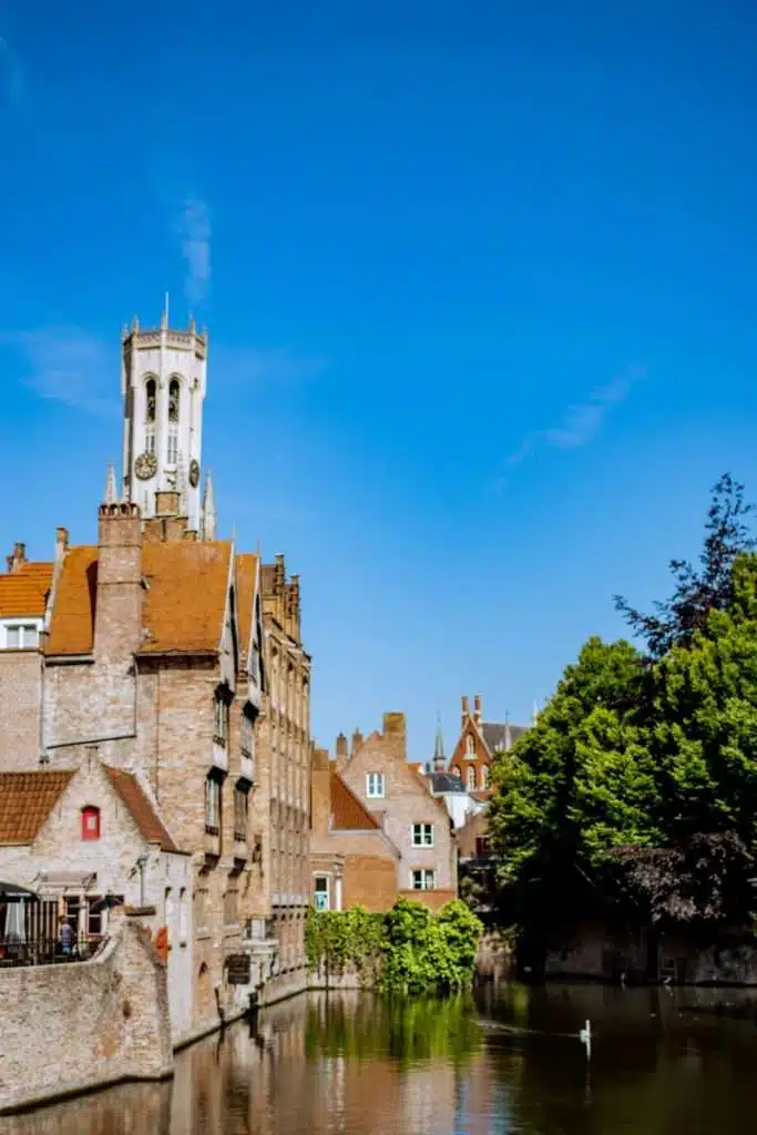 A scenic view of a historic European town with a canal in the foreground reveals one of the fun things to do in Bruges. The brick buildings boast red-tiled roofs, and a tall clock tower rises in the background. Lush trees line the canal under a clear blue sky.