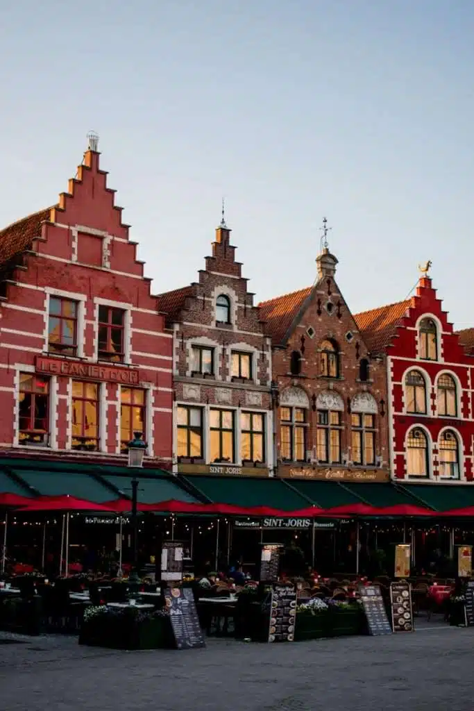 A row of colorful, historic gabled buildings in a located in Markt Square Bruges. The structures feature stepped facades and house cafes with outdoor seating. The sky is clear, adding a warm glow to the scene.