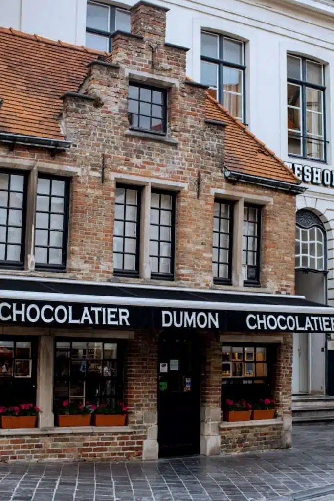 A charming brick building houses the Dumon Chocolatier shop. It features a steep, tiled roof and large front windows adorned with flower boxes. The black awning displays the shop's name in white lettering.
