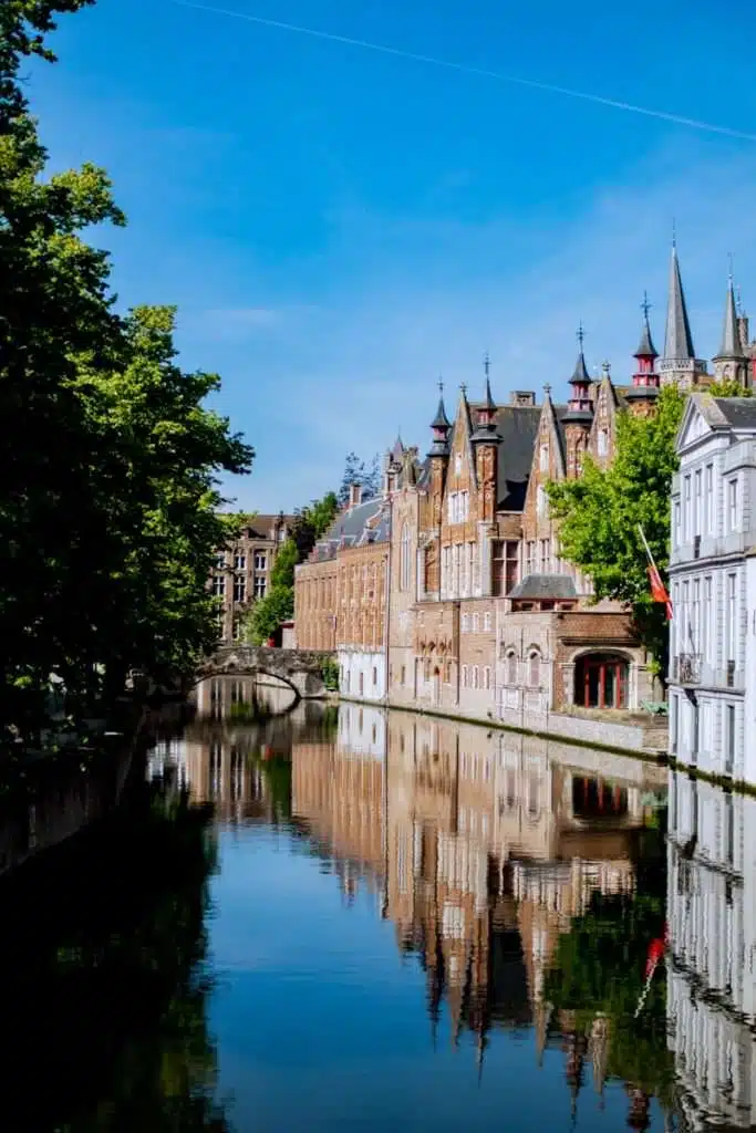 A scenic canal flanked by historic brick and stone buildings with pointed roofs in Bruges, Belgium. The calm water reflects the buildings and a small arched bridge in the background. Lush green trees line the canal under a clear blue sky.