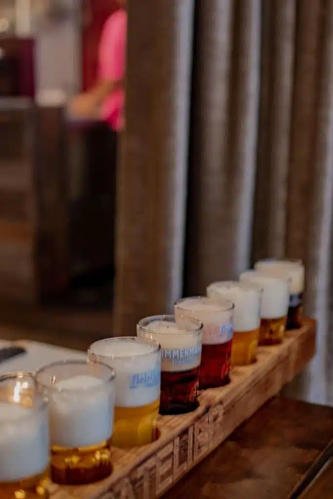 A wooden tray holds a row of beer glasses, each filled with brews in different shades and topped with foam—a taste of Bruges' vibrant beer scene. The softly blurred background hints at charming places to eat in Bruges, complementing the rich variety in the foreground.