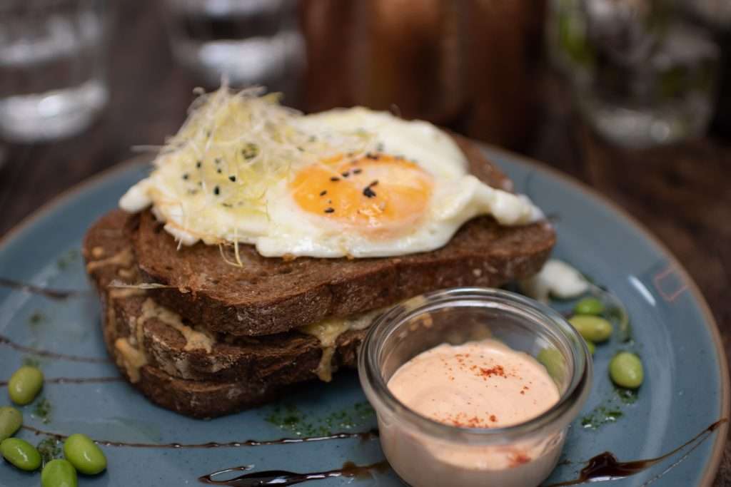 Two slices of toasted bread, crowned by fried eggs and garnished with sprouts and black sesame seeds. A small dish of creamy sauce sits beside scattered edamame beans.