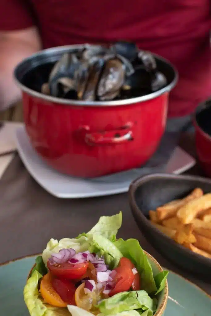 A dish of fresh salad with lettuce, cherry tomatoes, and diced red onions graces the foreground. Behind it, a pot of cooked mussels and a bowl of fries sit enticingly on a table—suggesting one of the cozy places to eat in Bruges. A person wearing a red shirt is seated at the table.