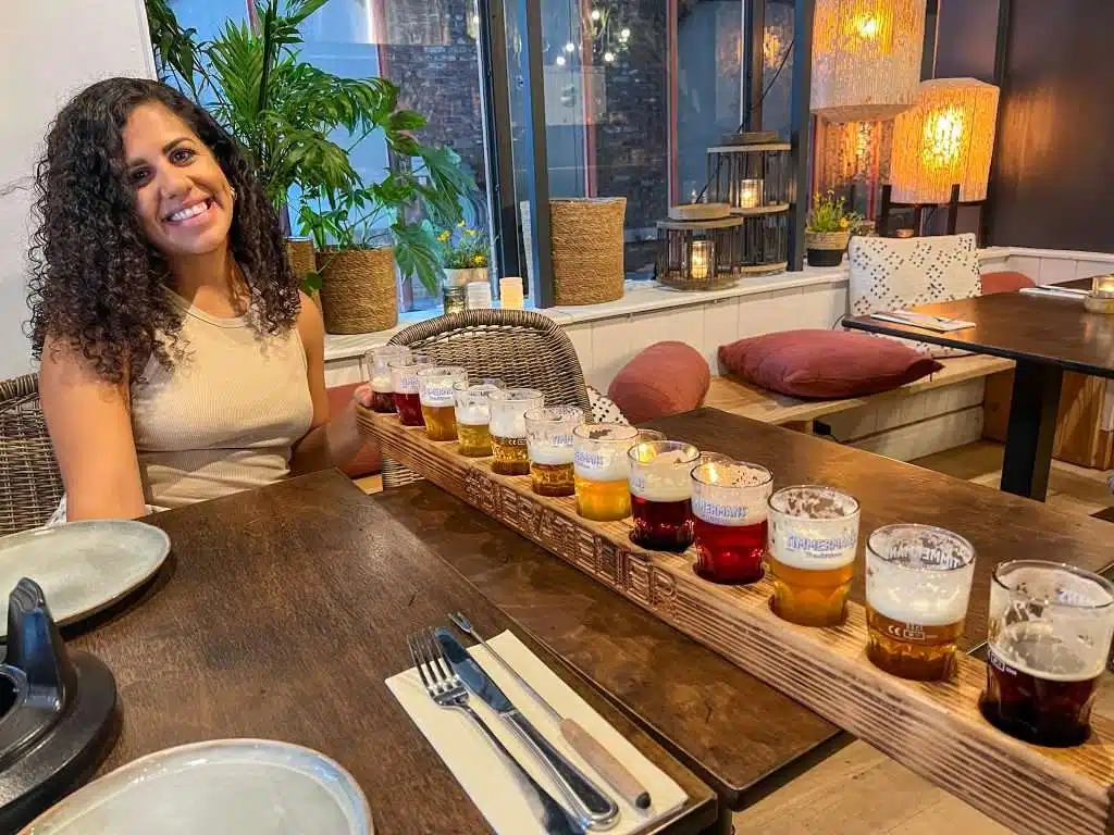 A woman with curly hair smiles while sitting at a wooden table in a cozy, modern restaurant in Bruges. In front of her is a long wooden tray with a variety of beers in small glasses. The background features plants and soft lighting—a perfect spot to explore amazing places to eat in Bruges.
