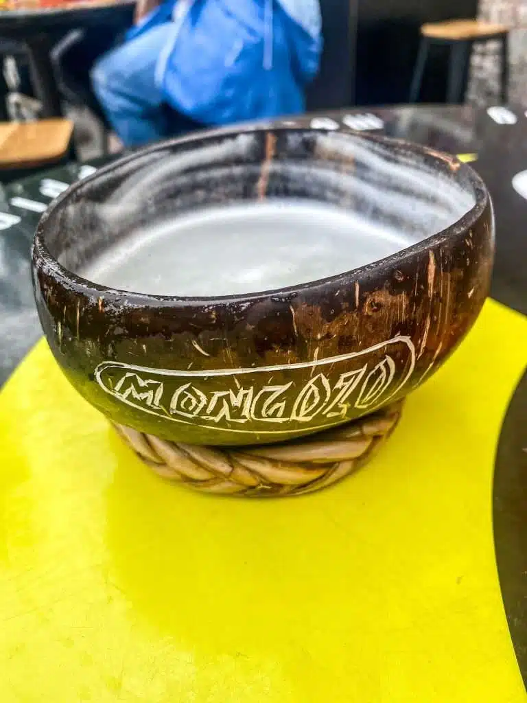 A wooden bowl with "MONGOZO" carved on the side, filled with a light liquid, evokes a taste of Bruges dining. The bowl rests on a yellow surface with a wicker base. In the blurred background, hints of outdoor furniture suggest charming places to eat in this picturesque city.