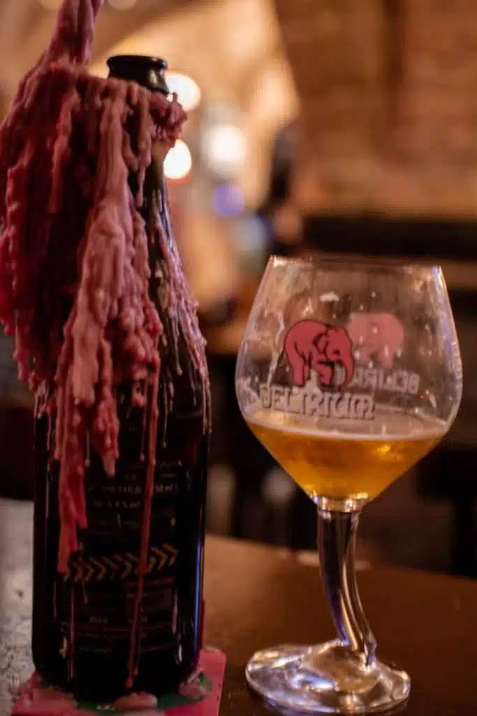 A bottle covered in melted wax sits next to a partially filled glass featuring a pink elephant logo and "Delirium" branding, evoking the cozy vibe of one of those charming places to eat in Bruges, nestled in a dimly lit bar or pub.