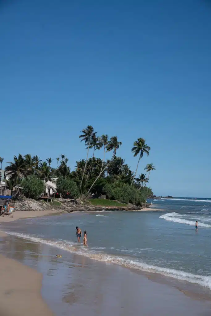 People stroll along a sandy beach with gentle waves under a clear blue sky in Ahangama. Palm trees and greenery line the shore, while a few buildings peek through—perfect for discovering top things to do in Ahangama.