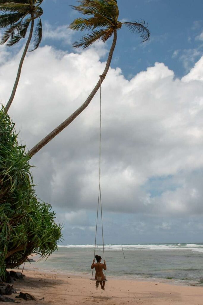 A person sits on a swing hanging from a tall, leaning palm tree on a sandy beach, facing the ocean under a partly cloudy skyโone of the unforgettable things to do in Ahangama.