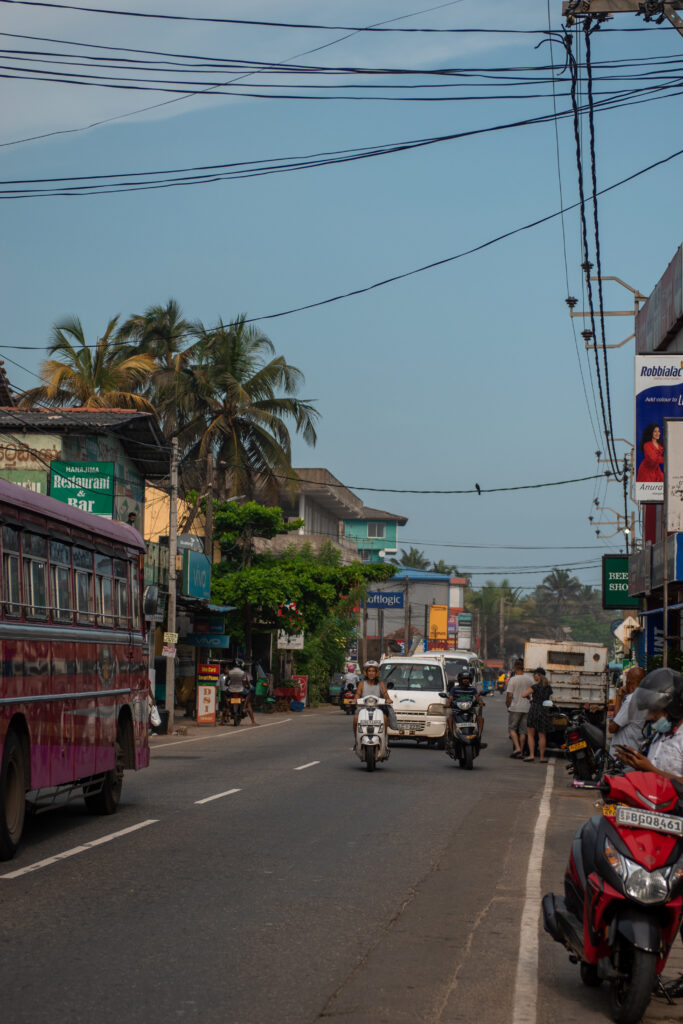 A busy street scene in India with cars, motorcycles, and a bus on the road, people walking on the sidewalk, shops and signboards lining both sidesโmuch like exploring local streets among the top things to do in Ahangama.