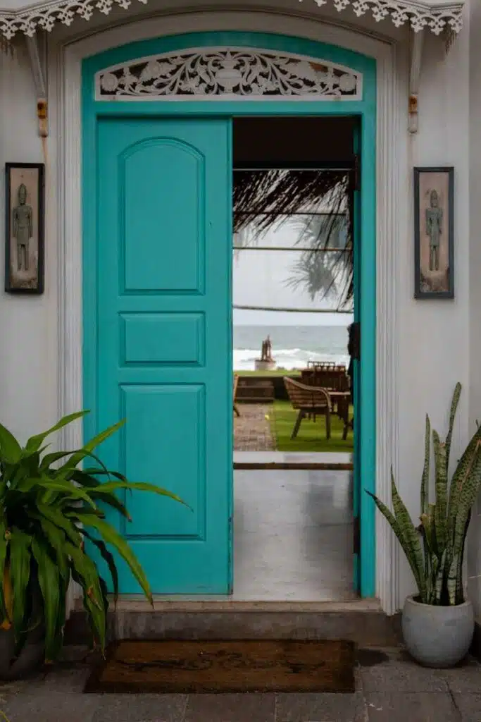 A turquoise door is partially open, revealing a patio with chairs, a table, palm trees, and the ocean in the background—an inviting scene that hints at relaxing things to do in Ahangama. Potted plants sit by the entrance.