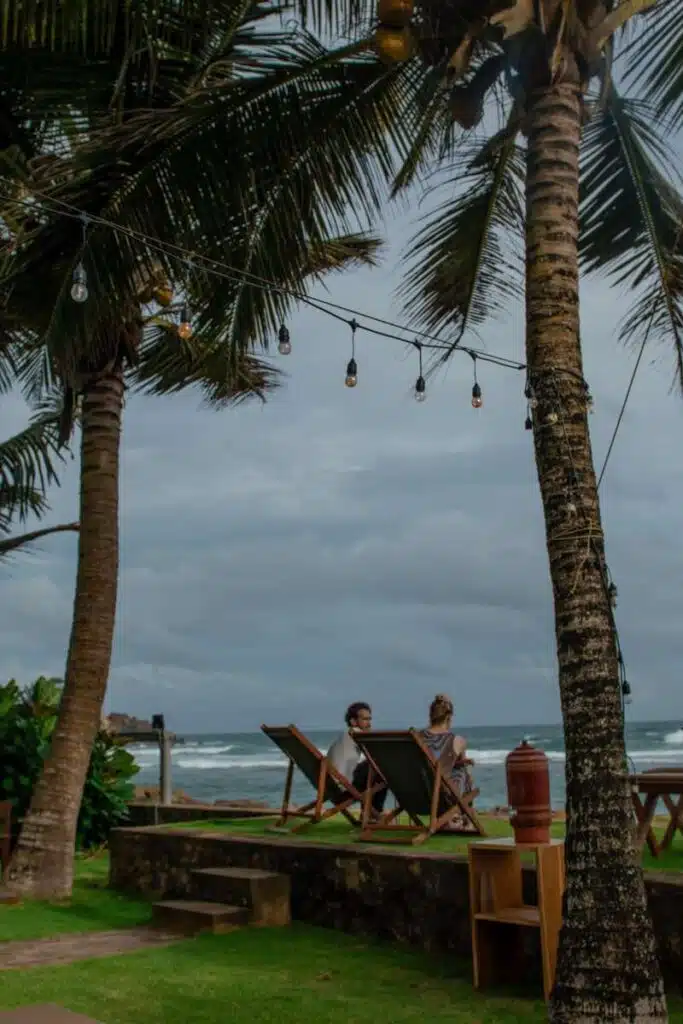 Two people relax on deck chairs by the ocean in Ahangama, surrounded by palm trees and string lights under a cloudy sky—a serene scene that captures the essence of things to do in Ahangama. Waves crash gently in the background.
