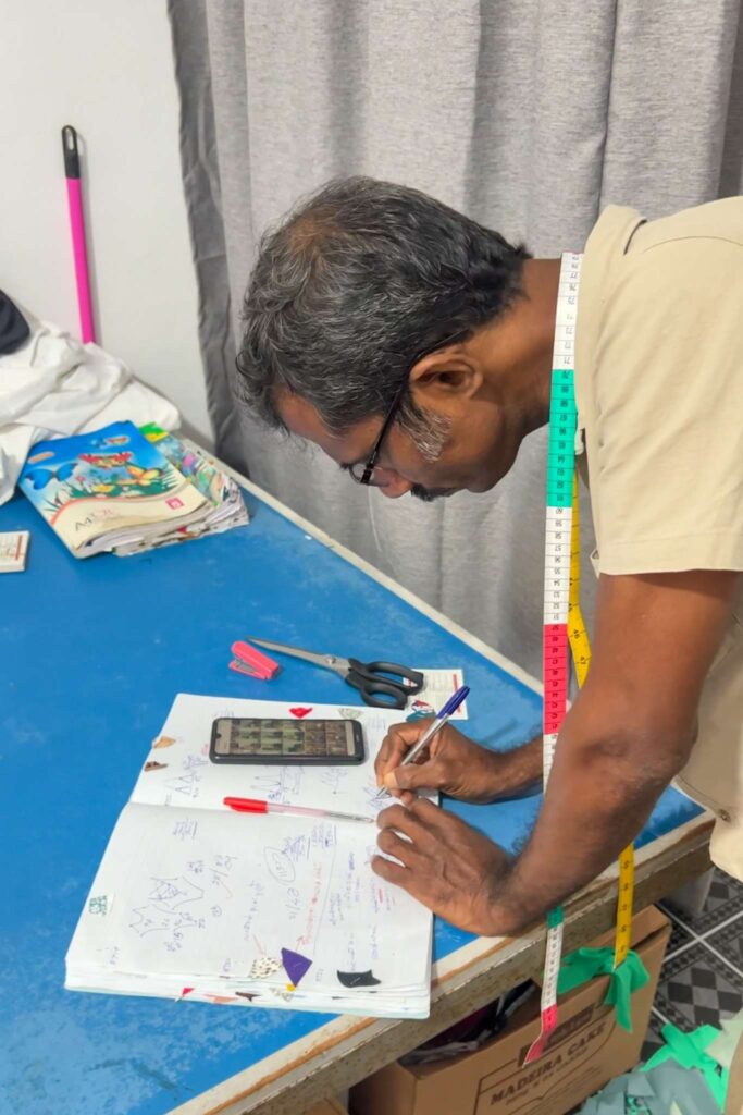 A man with glasses writes in a notebook on a table, with a smartphone and scissors nearby. Measuring tape hangs around his neck, and books and sewing tools are visible—perhaps planning creative things to do in Ahangama.