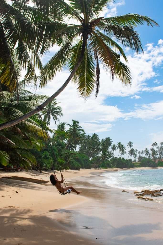 A person swings on a rope swing hanging from a tall palm tree over a sandy tropical beach—one of the must-try things to do in Hiriketiya—with lush greenery, swaying palms, and gentle waves under a partly cloudy sky.