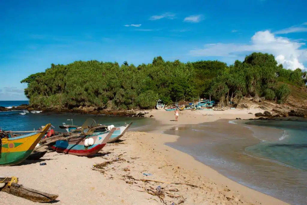 Several small, colorful fishing boats rest on a sandy beach near shallow, clear water—a perfect scene for those seeking Things To Do In Hiriketiya, with lush green trees and a small island in the background under a bright blue sky.
